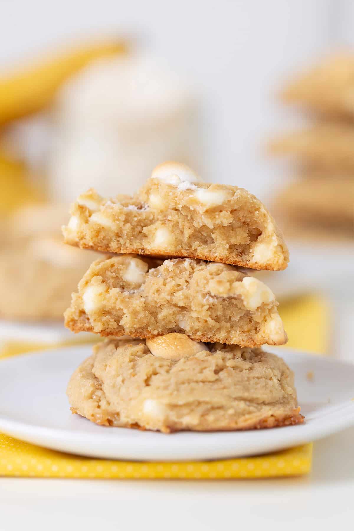 Three banana pudding cookies with white chocolate chips are stacked on a white plate; the top cookie is broken in half to reveal the soft, chewy texture and creamy chips inside.