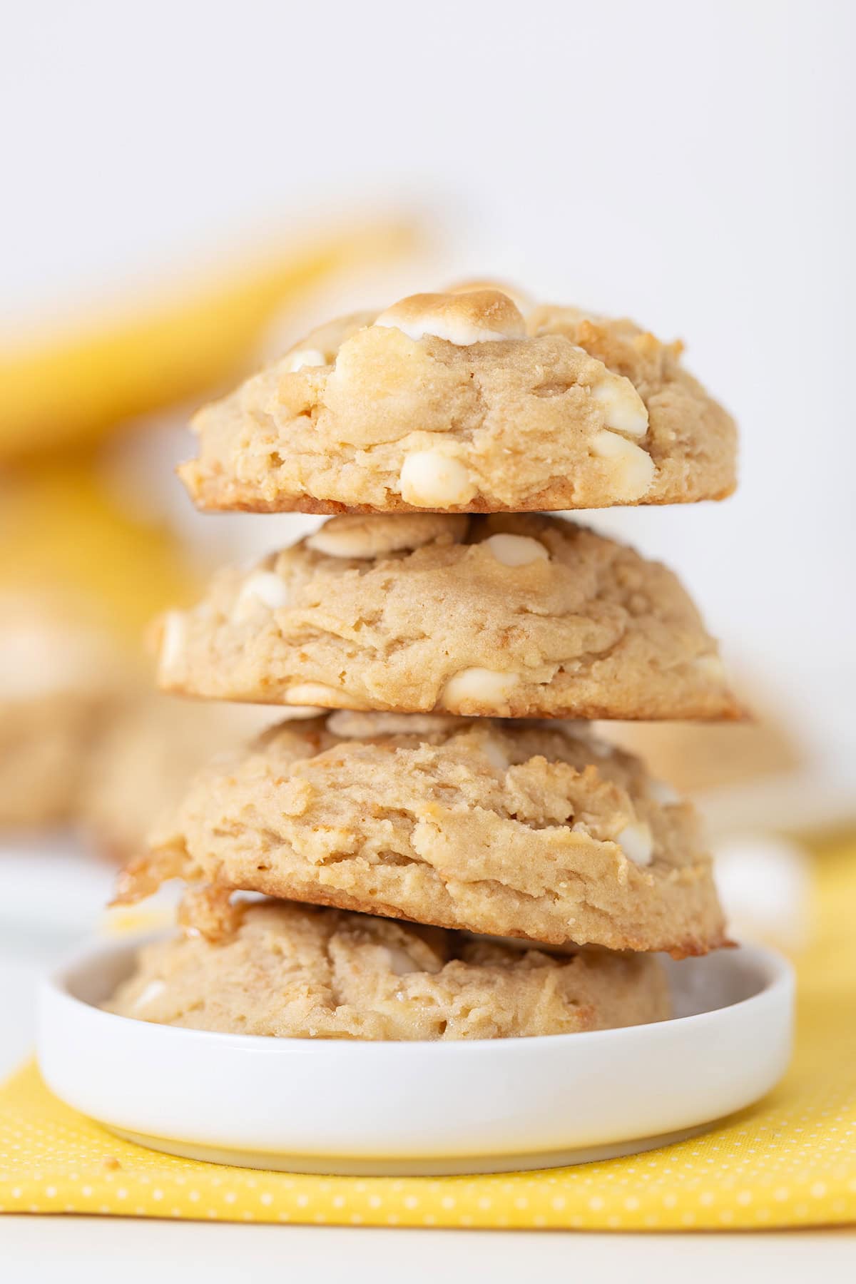A stack of three banana pudding cookies with white chocolate chips sits on a white plate, with a yellow napkin in the background.