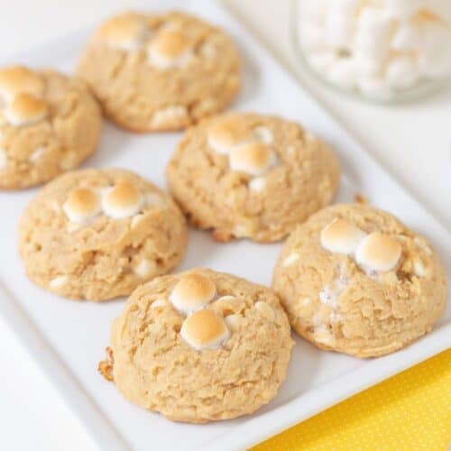 Six banana pudding cookies with toasted mini marshmallows and white chocolate chips on a white rectangular plate, with additional marshmallows in a glass bowl in the background.