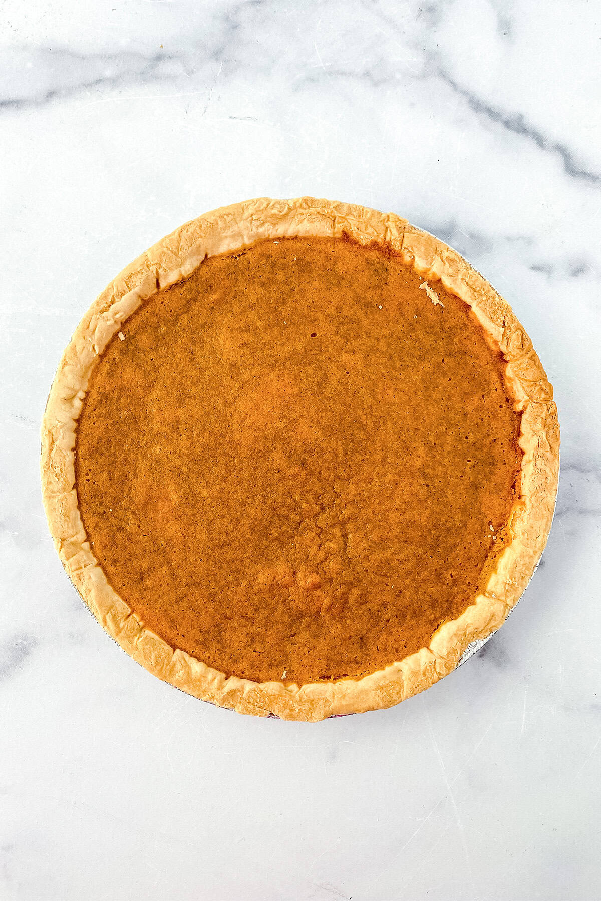 A whole sweet potato pie with a golden-brown filling and crust, featuring hints of browned butter, photographed from above on a white marble surface.