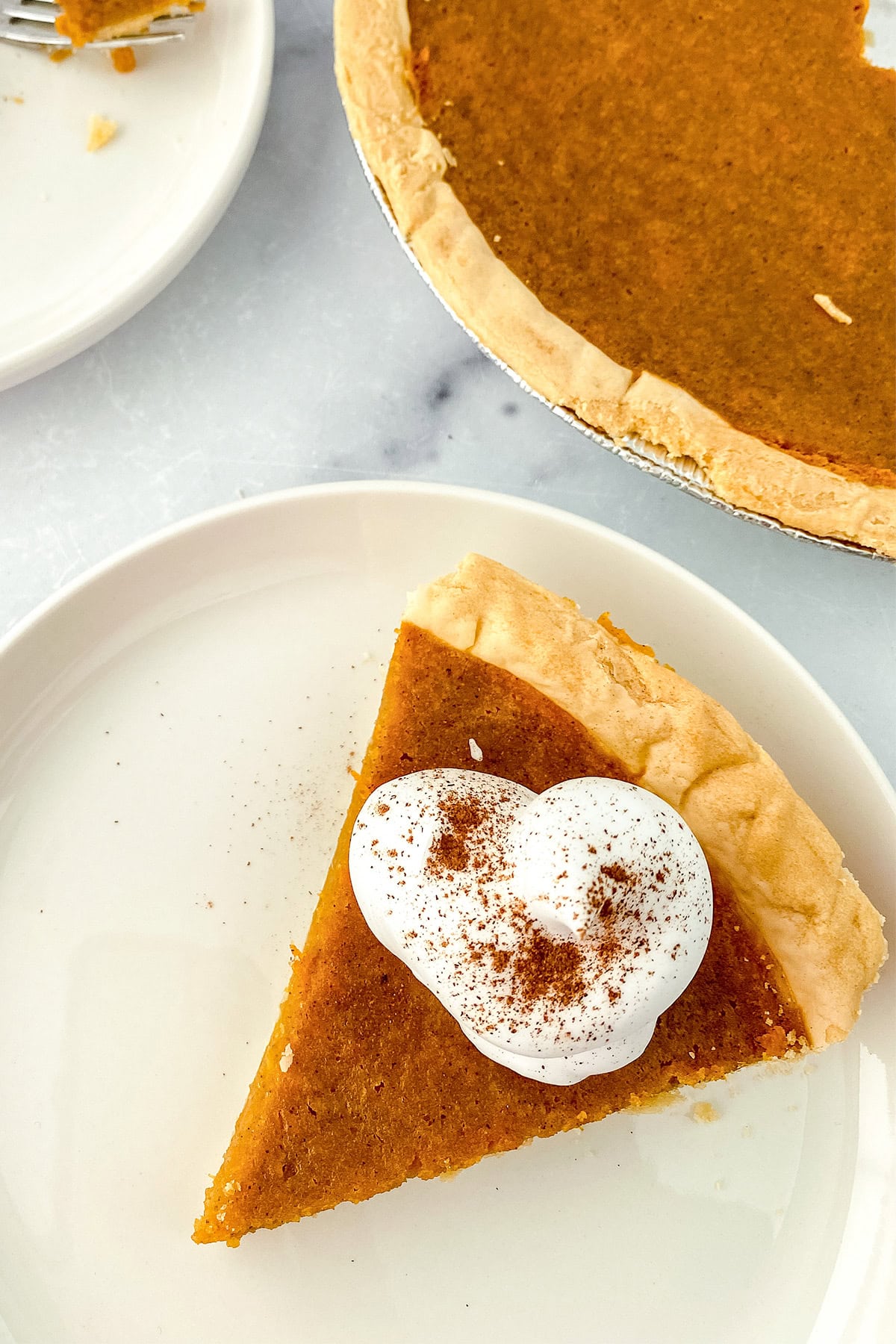 A slice of pumpkin pie topped with whipped cream and a sprinkle of cinnamon sits on a white plate next to the remaining pie, evoking the rich aroma of browned butter and cozy memories of homemade sweet potato pie.