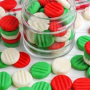 A glass jar filled with red, green, and white homemade butter mints, with more candies scattered on a white surface. A plaid cloth is in the background.