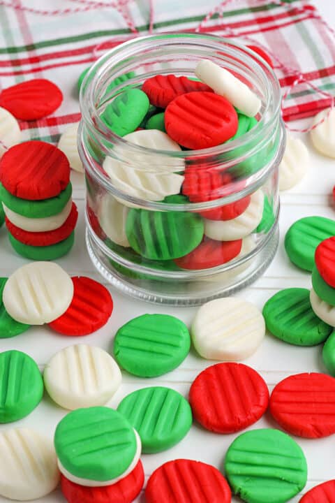 A glass jar filled with red, green, and white homemade butter mints, with more candies scattered on a white surface. A plaid cloth is in the background.