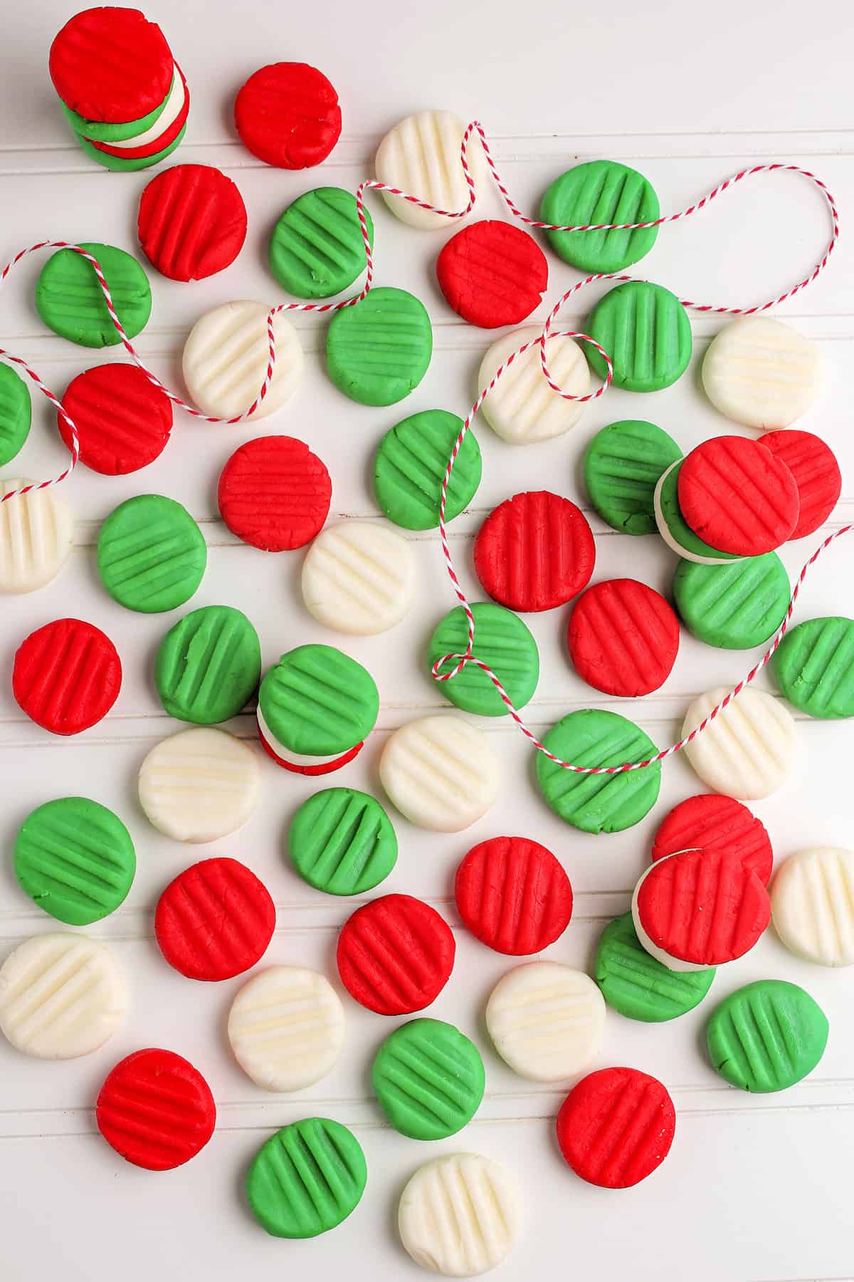 Red, green, and white Homemade Butter Mints with ridged tops are scattered on a white surface, some stacked together with a piece of red and white string among them.