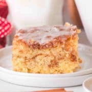 A square piece of Honey Bun Cake with icing on top sits on a white plate, with a cinnamon stick in the foreground.