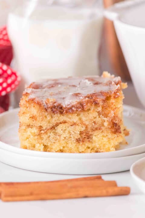 A square piece of Honey Bun Cake with icing on top sits on a white plate, with a cinnamon stick in the foreground.