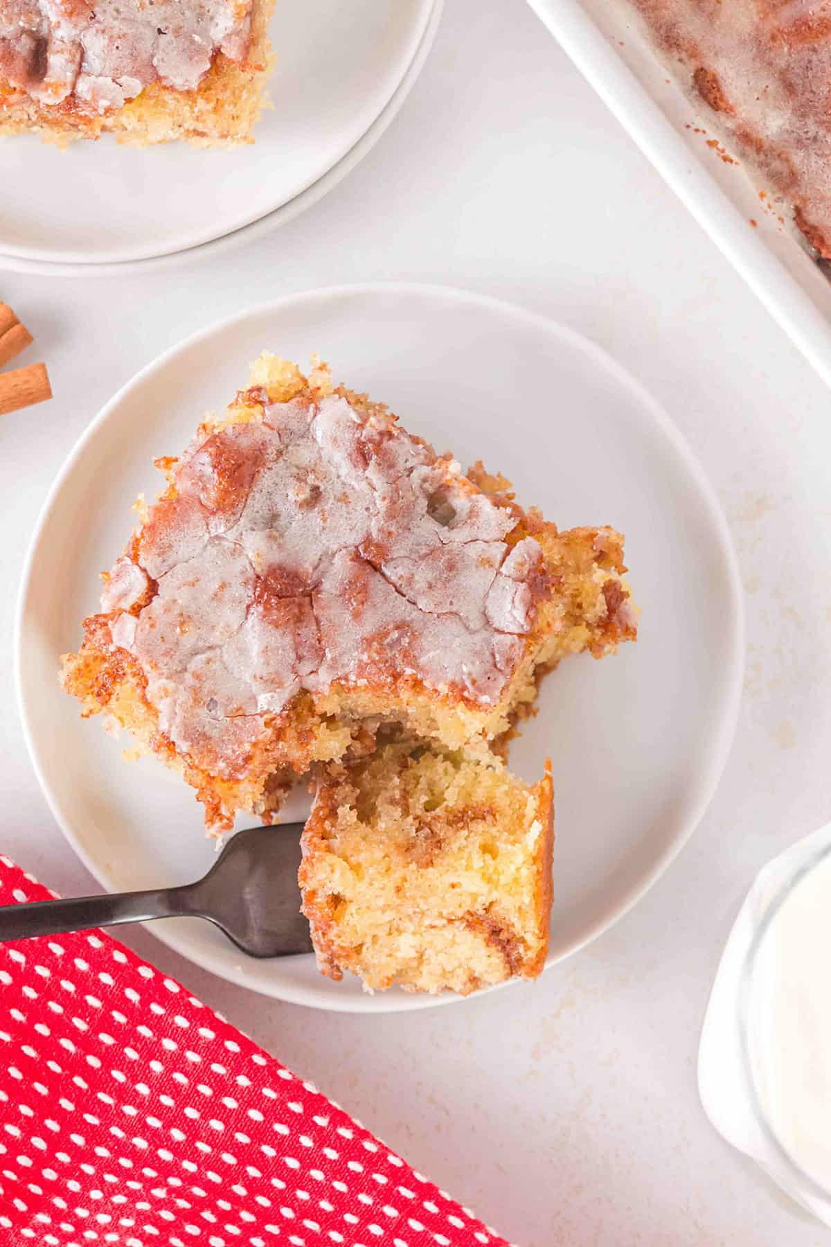 A slice of Honey Bun Cake with glaze sits on a white plate, a fork taking a bite, next to a red cloth and a cup of milk.