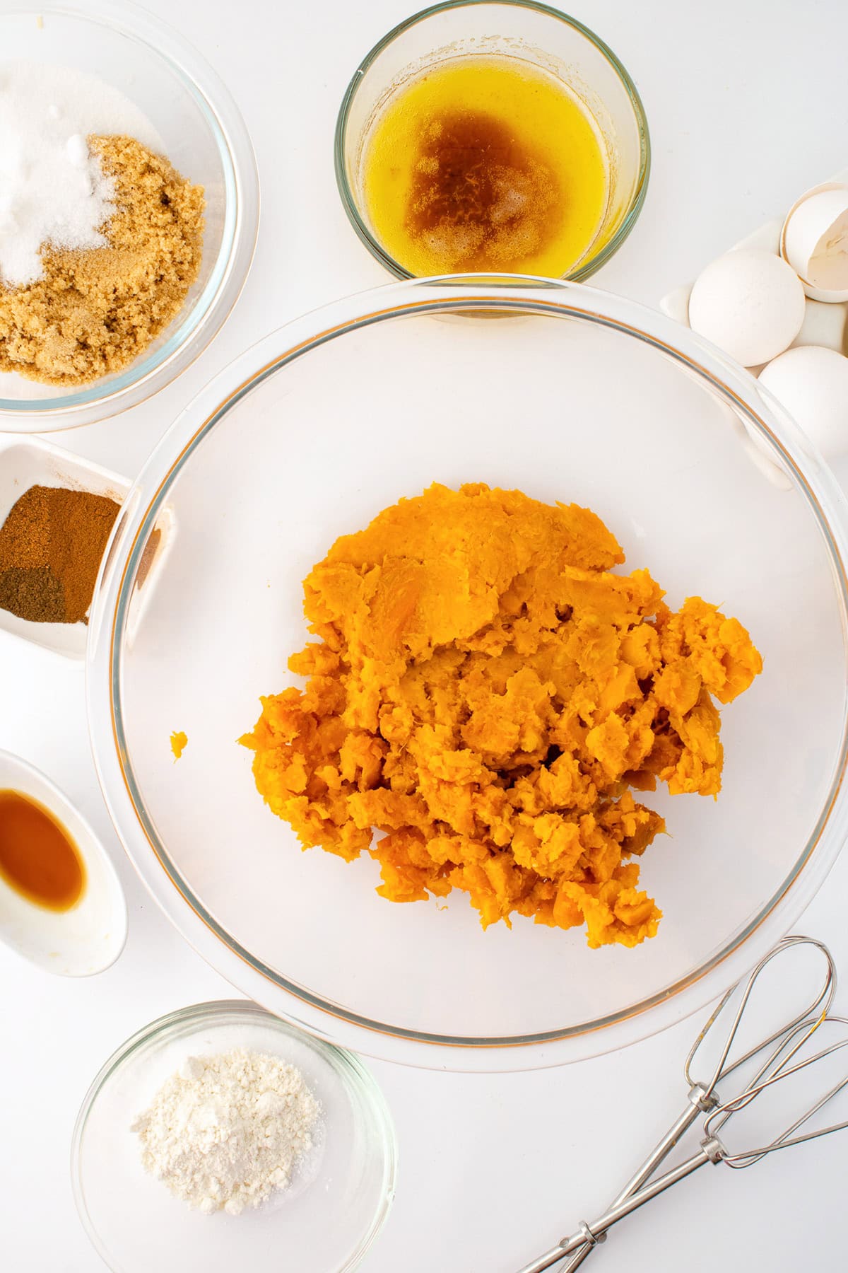 A glass bowl of mashed sweet potatoes for sweet potato pie is surrounded by bowls of brown sugar, browned butter, flour, vanilla extract, spices, eggs, and a hand mixer on a white surface.
