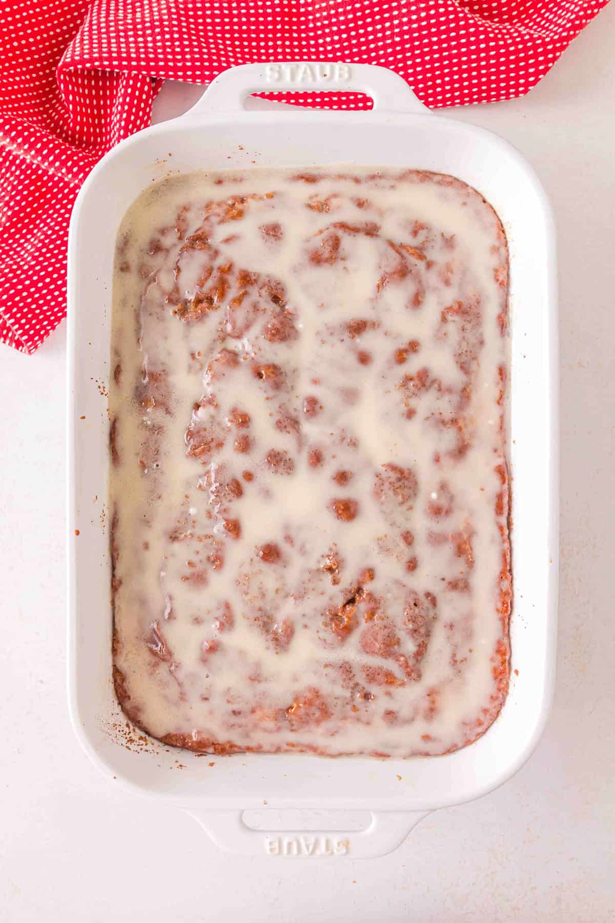 A rectangular white baking dish filled with Honey Bun Cake topped with a white glaze, placed on a white surface next to a red and white polka dot cloth.