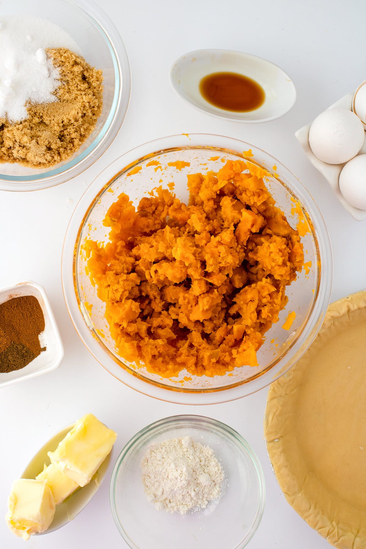 Overhead view of sweet potato pie ingredients, including mashed sweet potato, browned butter, sugar, eggs, pie crust, spices, flour, and vanilla extract on a white surface.