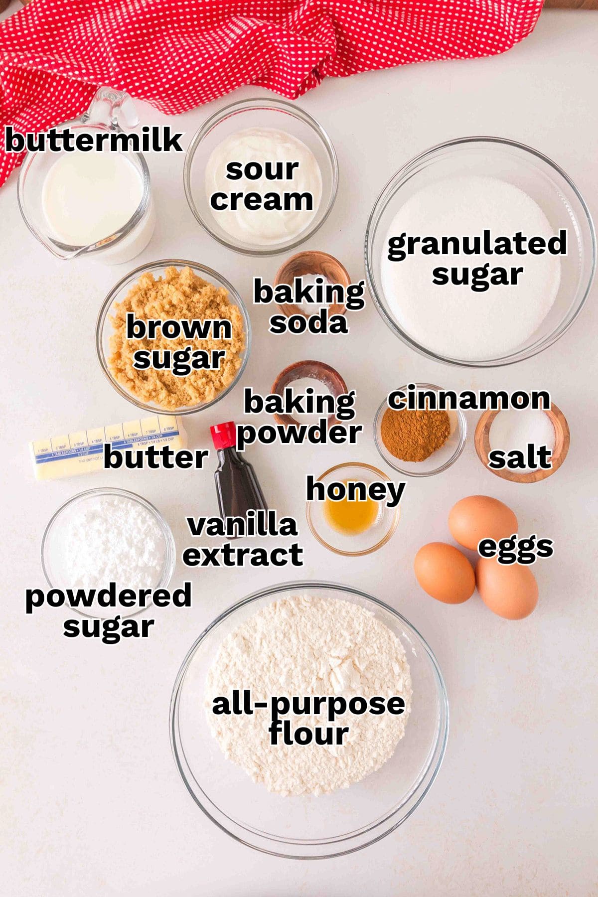 Various labeled baking ingredients, perfect for making a Honey Bun Cake, including buttermilk, sour cream, sugars, butter, baking soda and powder, cinnamon, salt, honey, vanilla extract, eggs, flour, and powdered sugar.