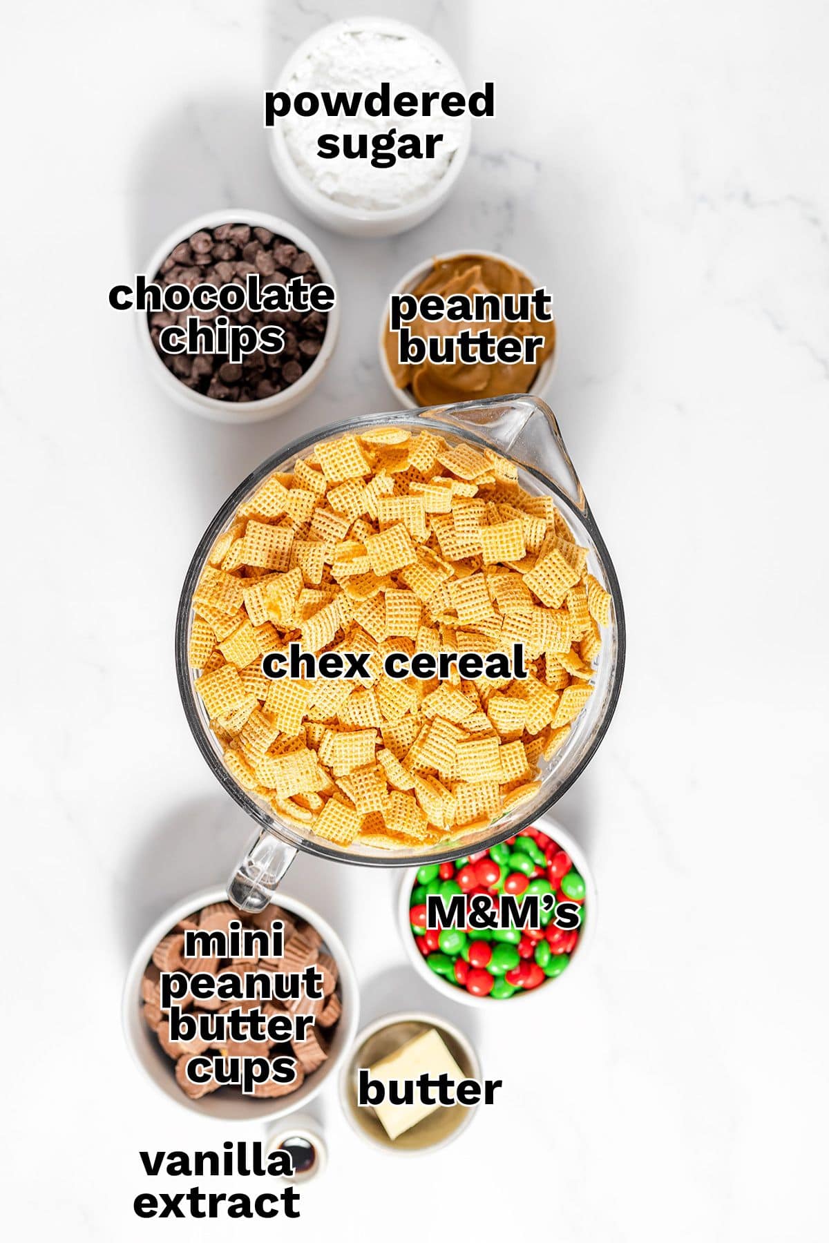 Bowls of ingredients labeled as powdered sugar, chocolate chips, peanut butter, Chex cereal, mini peanut butter cups, M&M's, butter, and vanilla extract arranged on a white surface—perfect for making Christmas Puppy Chow or Reindeer Food.