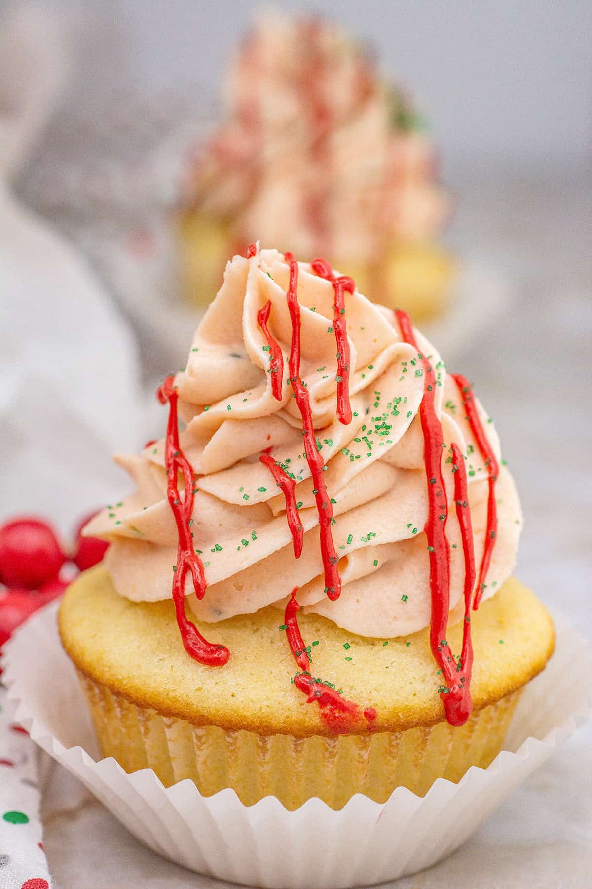 A vanilla cupcake with pink frosting, red drizzle, and green sprinkles, reminiscent of Christmas Tree Cupcakes, sits in a white paper liner on a light surface.