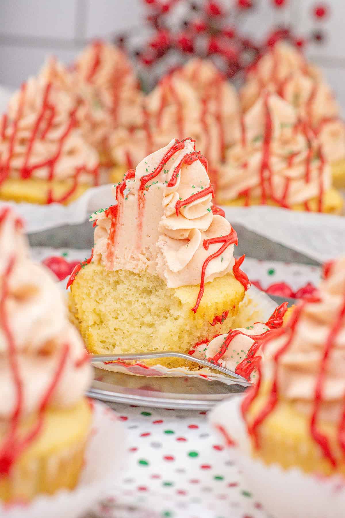 A yellow cupcake with pink swirled frosting and red drizzle, inspired by Little Debbie Christmas Tree Cupcakes, sits on a plate with a bite taken out, surrounded by similar cupcakes on a dotted tablecloth.
