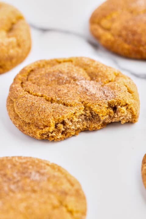 A close-up of a round Pumpkin Snickerdoodle cookie with a bite taken out, covered in cinnamon sugar, surrounded by other whole cookies on a white surface—a perfect treat for fans of fall desserts.