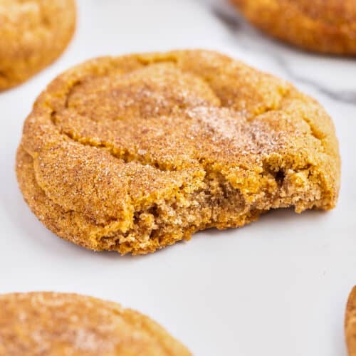 A close-up of a round Pumpkin Snickerdoodle cookie with a bite taken out, covered in cinnamon sugar, surrounded by other whole cookies on a white surface—a perfect treat for fans of fall desserts.