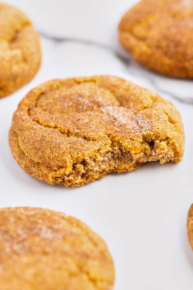 A close-up of a round Pumpkin Snickerdoodle cookie with a bite taken out, covered in cinnamon sugar, surrounded by other whole cookies on a white surface—a perfect treat for fans of fall desserts.