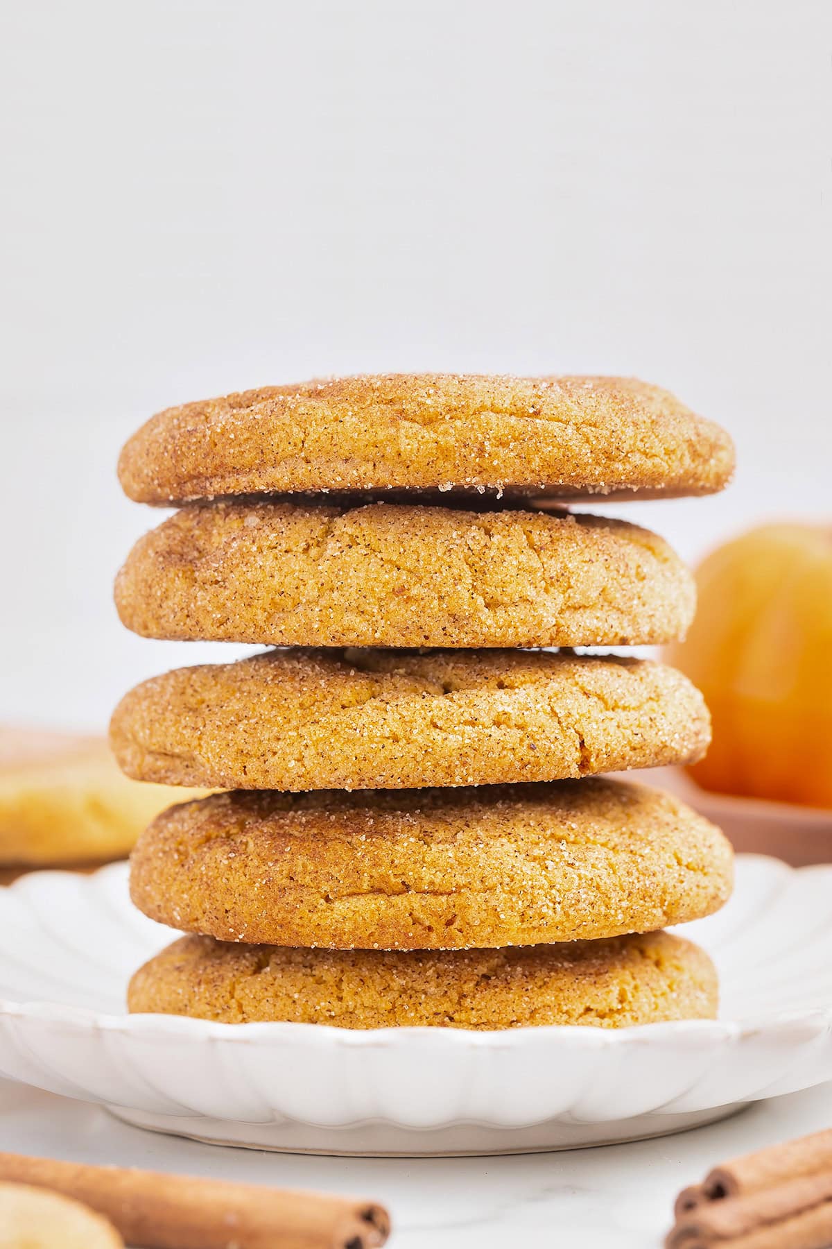 A stack of five round, golden-brown Pumpkin Snickerdoodles is arranged on a white plate with a blurred background.