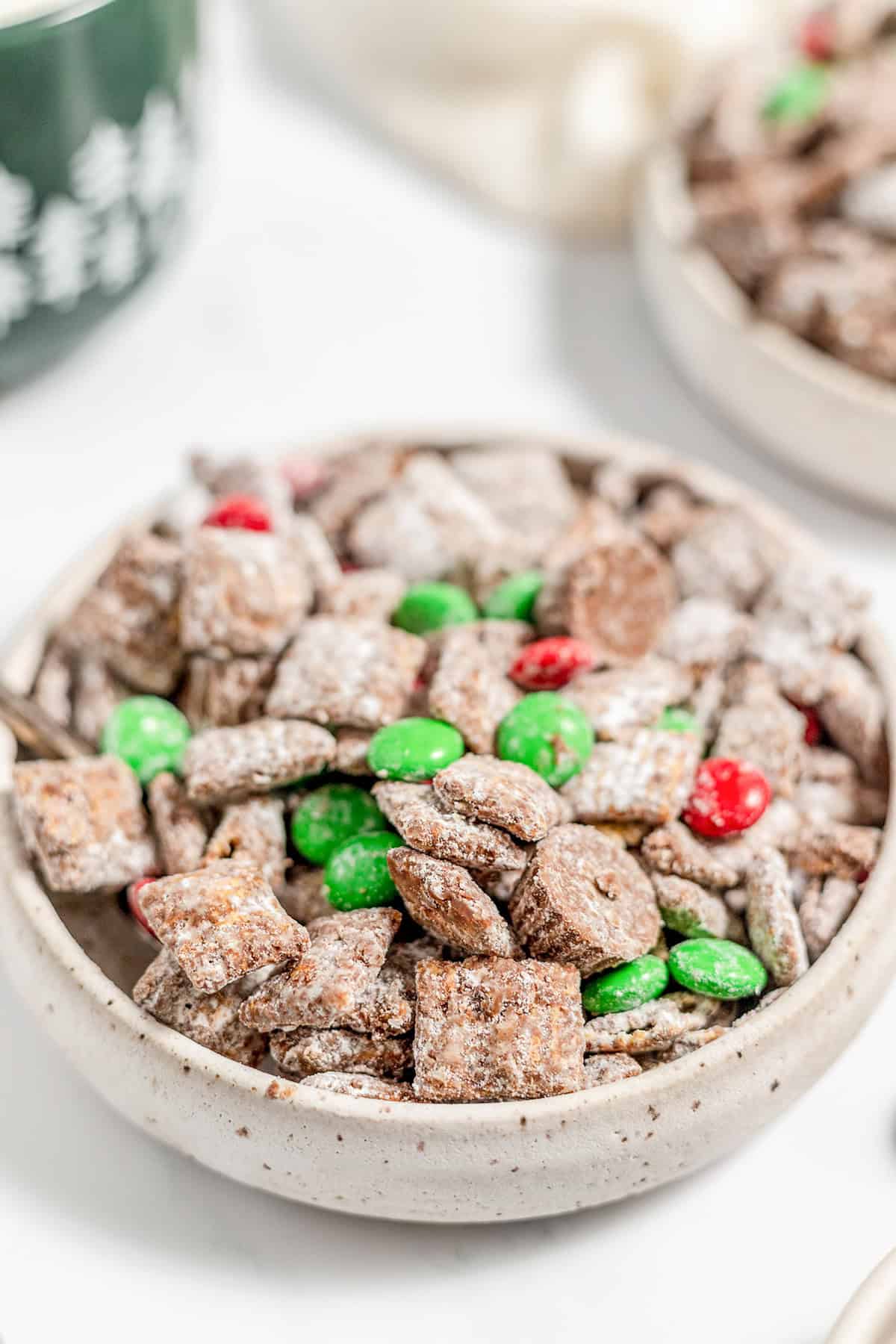 A bowl of Christmas Puppy Chow features chocolate-coated cereal squares dusted with powdered sugar and mixed with festive red and green candy-coated chocolates.
