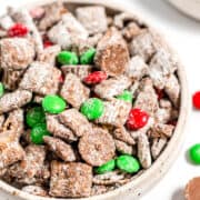 A bowl of Christmas Puppy Chow, a festive chocolate-coated cereal snack mix with powdered sugar, green and red candy-coated chocolates, and chocolate pieces—perfect as a Christmas snack or Reindeer Food!.