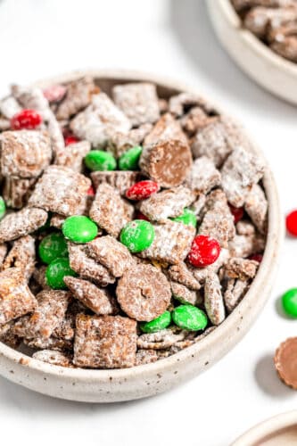 A bowl of Christmas Puppy Chow, a festive chocolate-coated cereal snack mix with powdered sugar, green and red candy-coated chocolates, and chocolate pieces—perfect as a Christmas snack or Reindeer Food!.