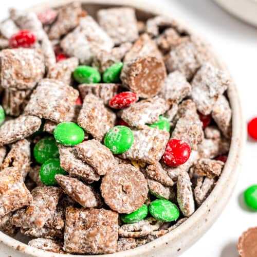A bowl of Christmas Puppy Chow, a festive chocolate-coated cereal snack mix with powdered sugar, green and red candy-coated chocolates, and chocolate pieces—perfect as a Christmas snack or Reindeer Food!.