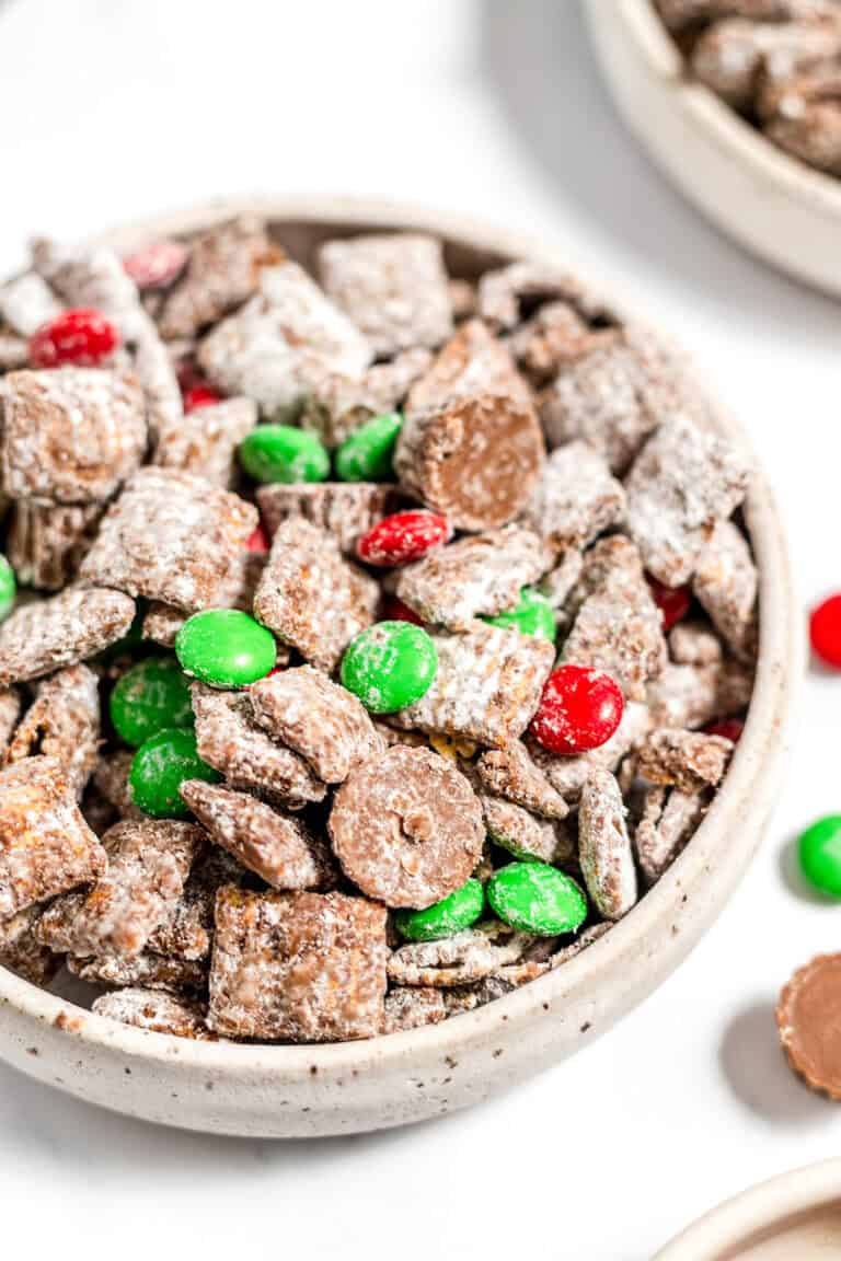 A bowl of Christmas Puppy Chow, a festive chocolate-coated cereal snack mix with powdered sugar, green and red candy-coated chocolates, and chocolate pieces—perfect as a Christmas snack or Reindeer Food!.