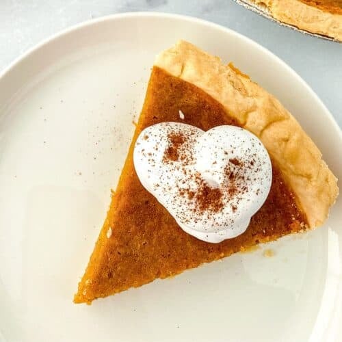 A slice of pumpkin pie topped with a dollop of whipped cream and a sprinkle of cinnamon, served on a white plate with hints of browned butter reminiscent of classic sweet potato pie.