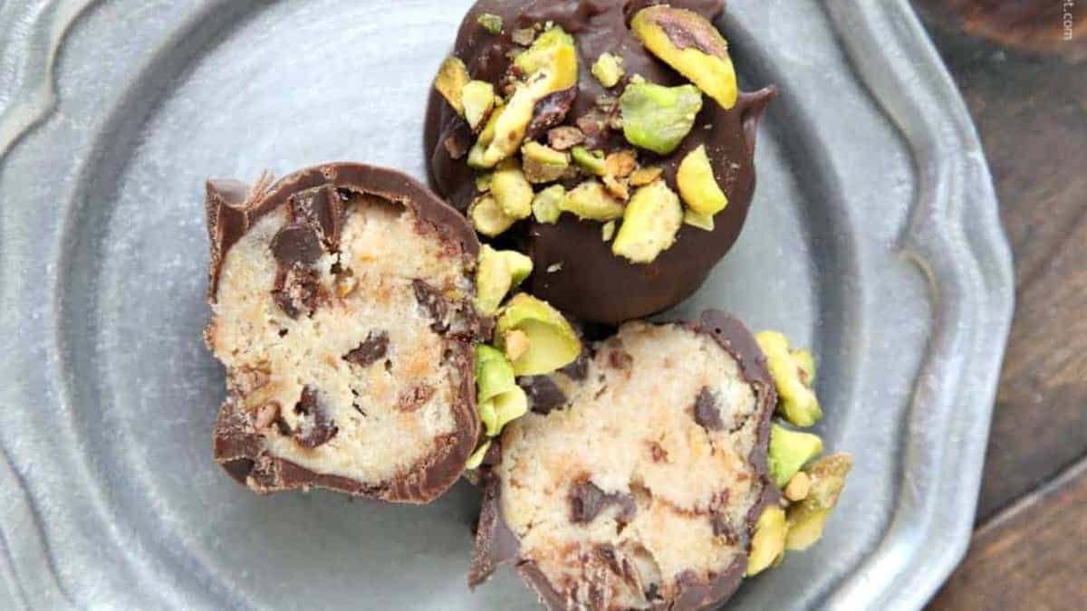 Three chocolate-coated homemade candies, two cut open to reveal a cookie dough-like filling with chocolate chips, topped with chopped pistachios, perfect for gifting in Christmas cookie boxes on a silver plate.