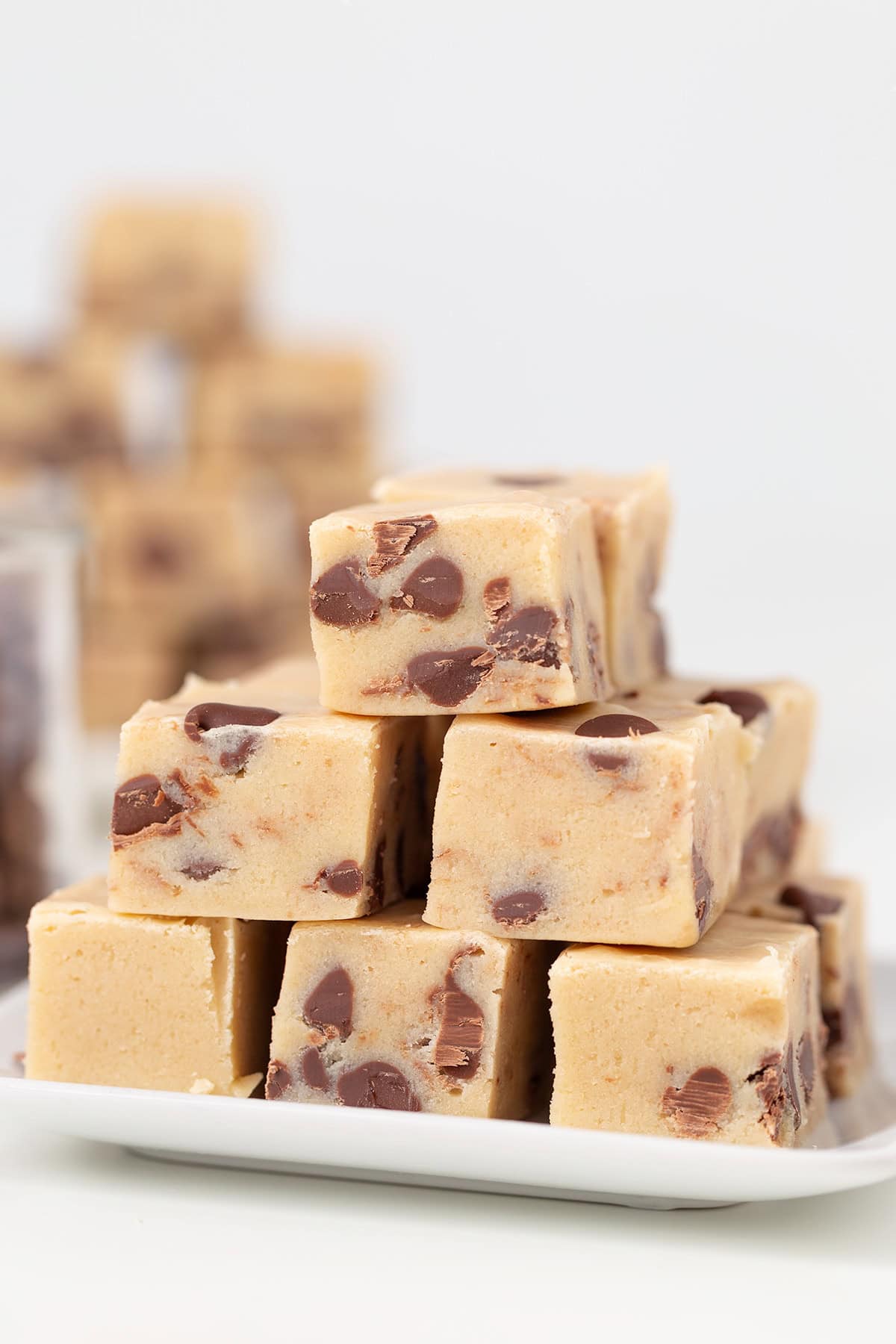 A stack of rich Cookie Dough Fudge squares sits on a white plate, with extra pieces of the creamy treat scattered invitingly in the background.