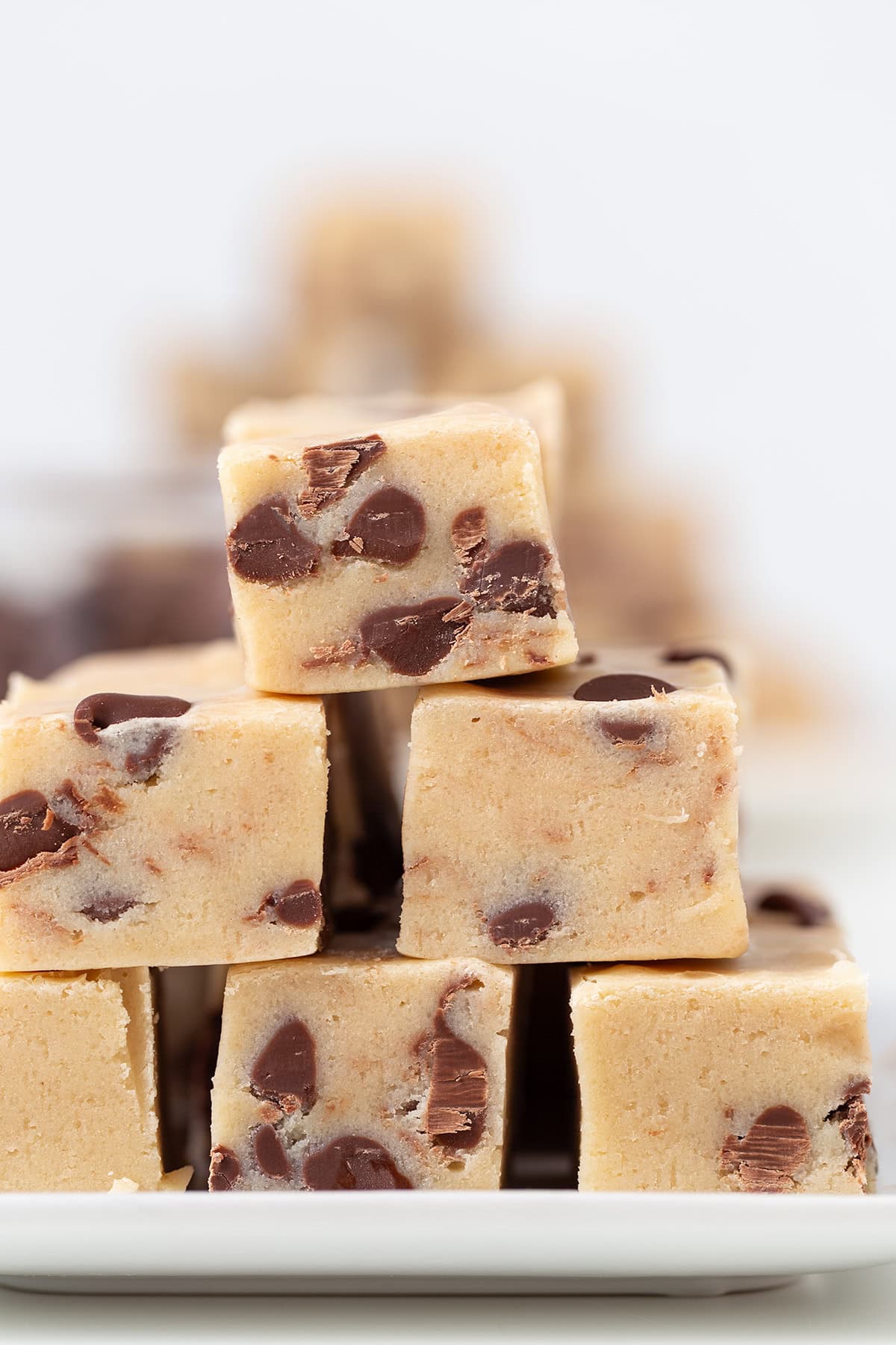 A close-up of stacked squares of cookie dough fudge, loaded with visible chocolate chips and arranged on a white plate.