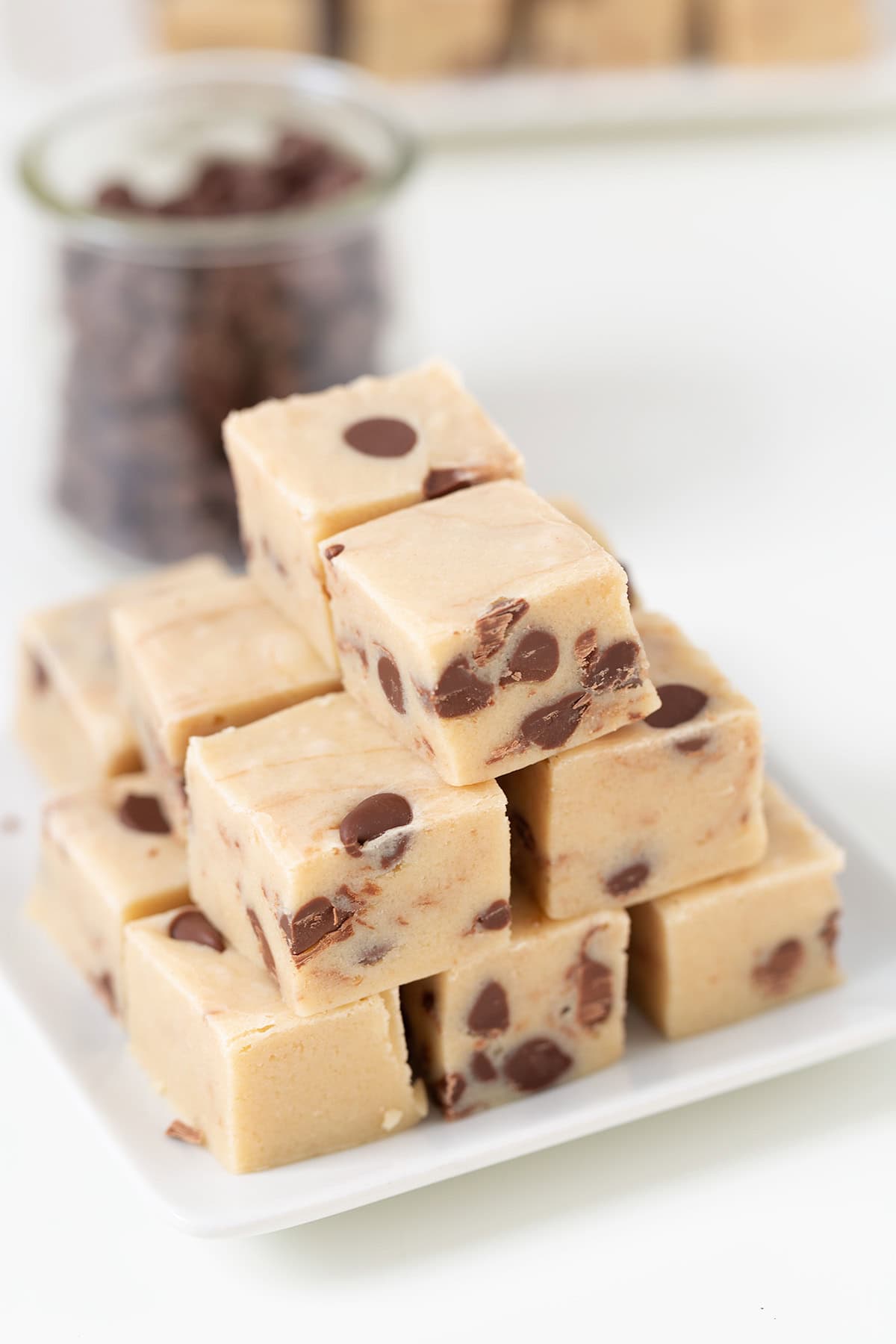 A stack of square pieces of cookie dough fudge with chocolate chips sits on a white plate, while a glass jar filled with extra chocolate chips adds charm in the background.