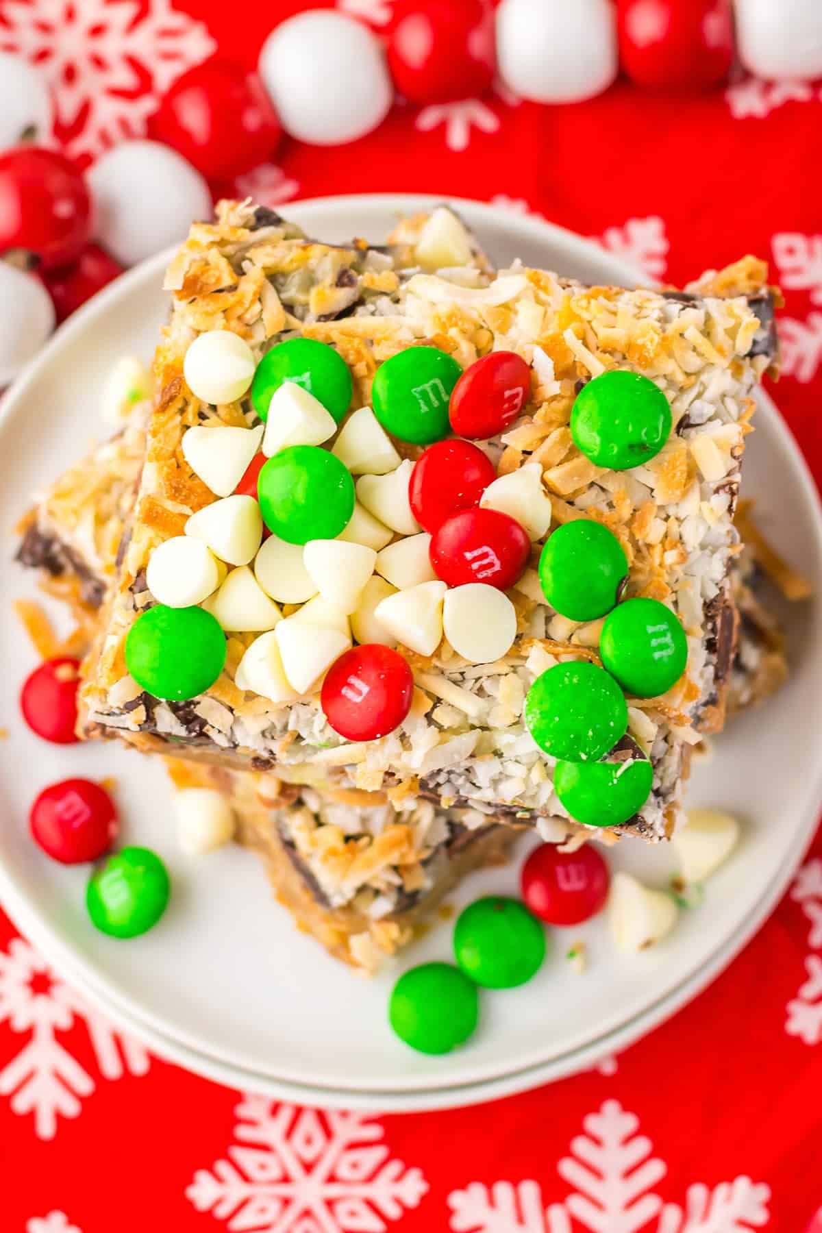 A plate of Christmas Magic Cookie Bars topped with white chocolate chips, red and green candies, and coconut flakes, set on a red snowflake-patterned tablecloth.