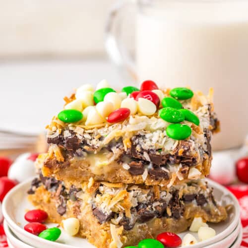 Two Christmas Magic Cookie Bars topped with chocolate chips, shredded coconut, white chocolate chips, and red and green candies are stacked on a plate with a glass of milk in the background.