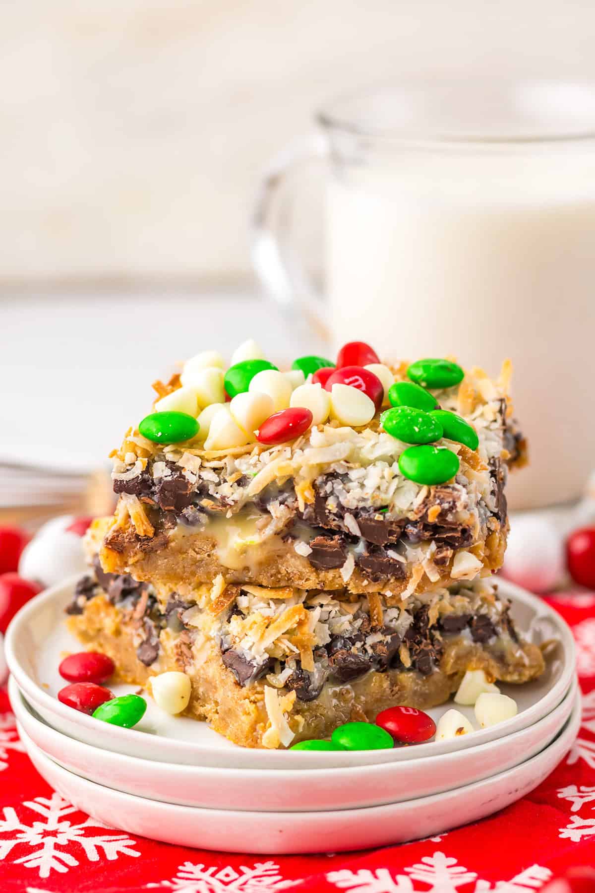Two Christmas Magic Cookie Bars topped with chocolate chips, shredded coconut, white chocolate chips, and red and green candies are stacked on a plate with a glass of milk in the background.