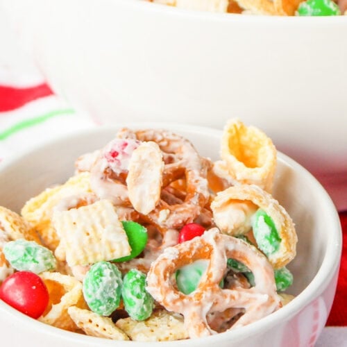A white bowl filled with a festive Christmas Snack Mix of pretzels, square cereals, candy-coated chocolates, and other coated treats. Another larger bowl is in the background—perfect for sharing this Christmas White Trash Recipe.