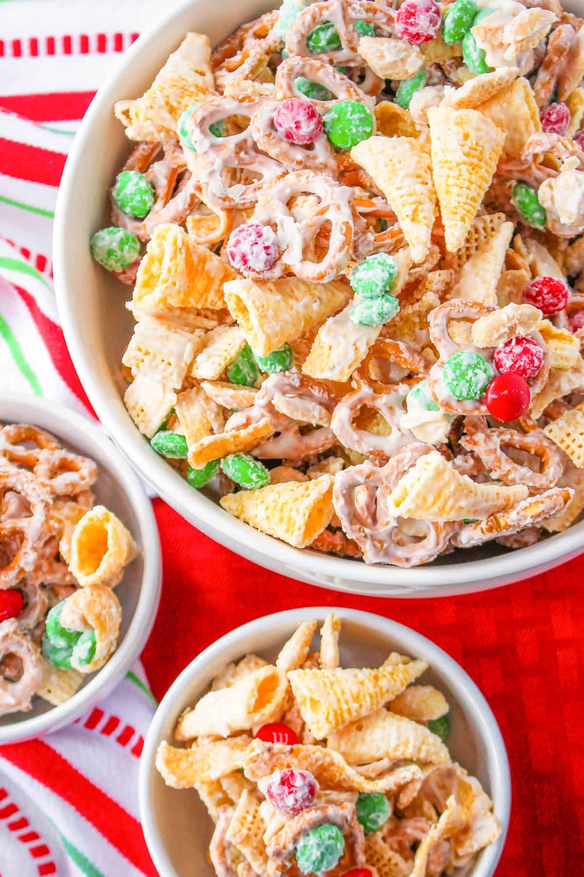 A large bowl and two small bowls filled with a Christmas White Trash Recipe snack mix of Bugles, pretzels, green and red candies, and cereal pieces all coated in white chocolate.