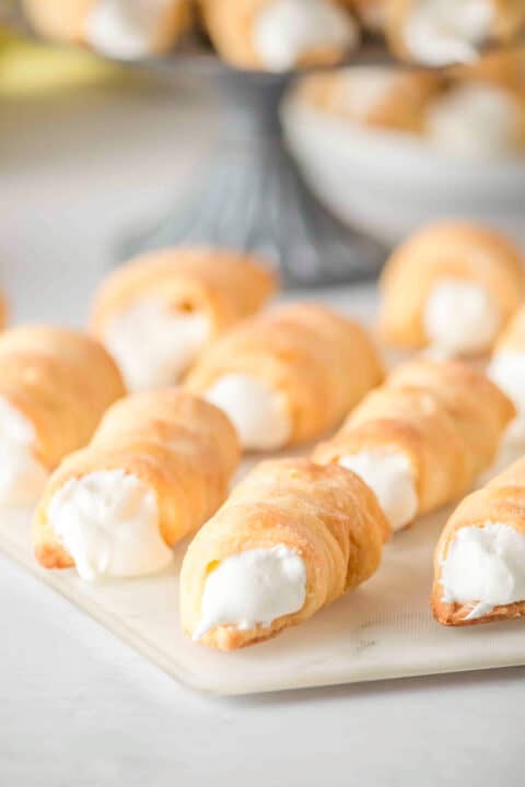 Several cream-filled Lady Locks pastry horns are arranged on a white surface, with more delicious cookies visible in the blurred background.