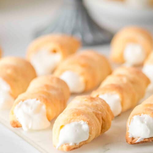 Several cream-filled Lady Locks pastry horns are arranged on a white surface, with more delicious cookies visible in the blurred background.