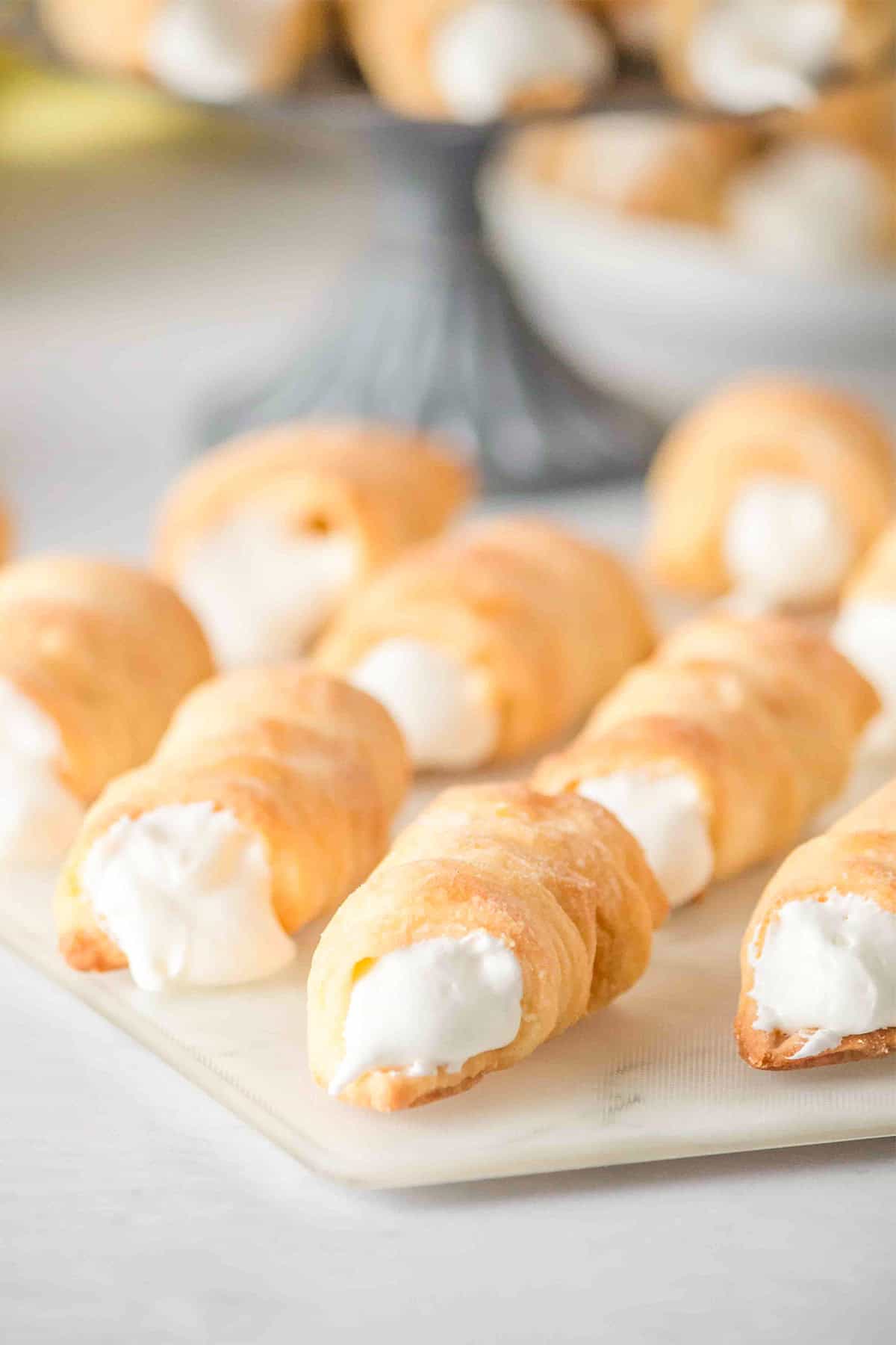 Several cream-filled Lady Locks pastry horns are arranged on a white surface, with more delicious cookies visible in the blurred background.
