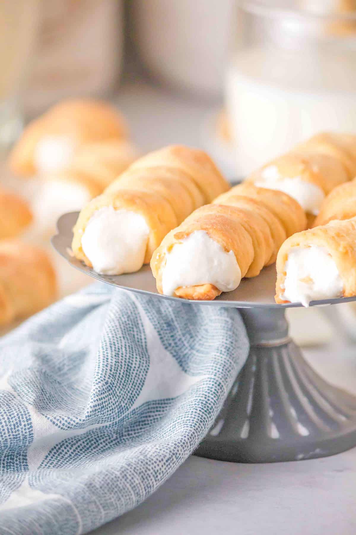 A grey cake stand holds several cream-filled Lady Locks pastry horns, with a blue and white cloth draped beneath.