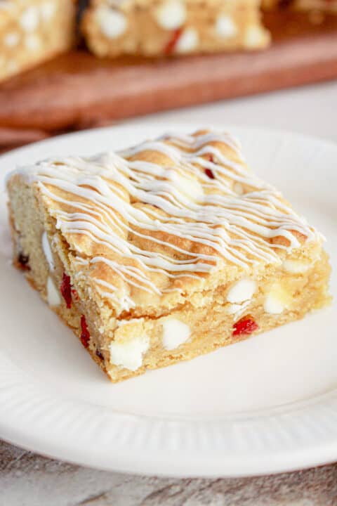 A blondie dessert bar with white chocolate chips, cranberry bits, and a white icing drizzle on top, served on a white plate.