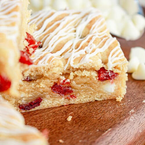 A close-up of a cranberry blondie bar with white chocolate chips and red fruit pieces, drizzled with white icing, on a wooden board. White chocolate chips are scattered in the background.