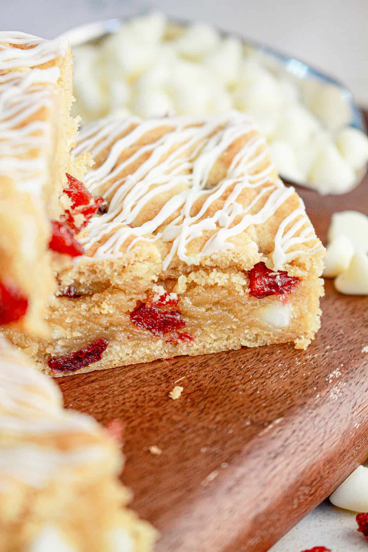 A close-up of a cranberry blondie bar with white chocolate chips and red fruit pieces, drizzled with white icing, on a wooden board. White chocolate chips are scattered in the background.