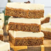 Three square slices of frosted Gingerbread Cookie Bars are stacked on a wooden board, with more pieces and plates visible in the background.