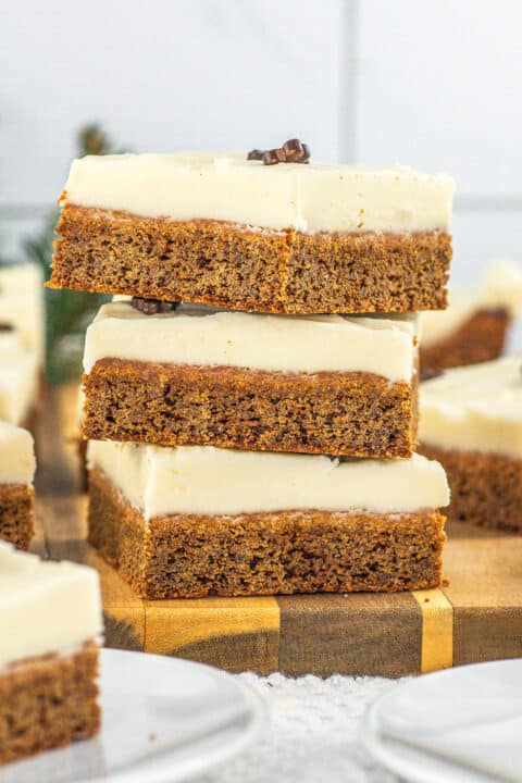 Three square slices of frosted Gingerbread Cookie Bars are stacked on a wooden board, with more pieces and plates visible in the background.