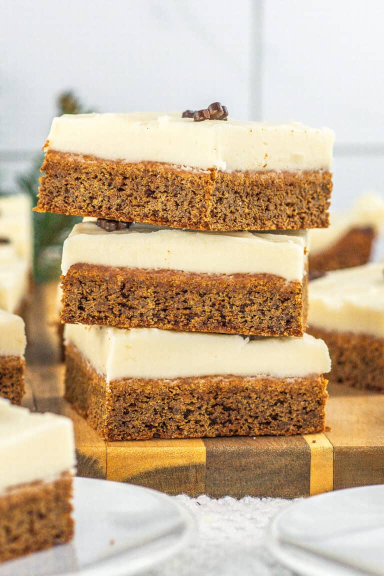 Three square slices of frosted Gingerbread Cookie Bars are stacked on a wooden board, with more pieces and plates visible in the background.