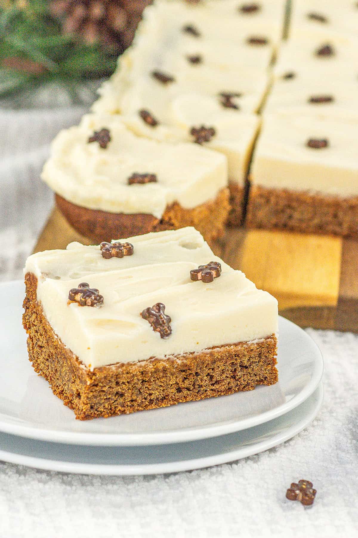 A frosted Gingerbread Cookie Bar topped with small chocolate decorations sits on a white plate, with more bars and a wooden board in the background.