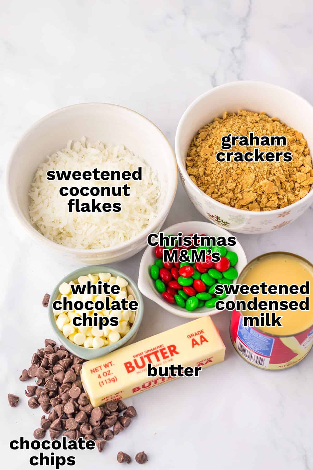 Bowls and containers with labeled ingredients for Christmas Magic Cookie Bars: sweetened coconut flakes, graham crackers, white chocolate chips, Christmas M&M’s, sweetened condensed milk, chocolate chips, and butter. Perfect for festive Christmas cookies!.