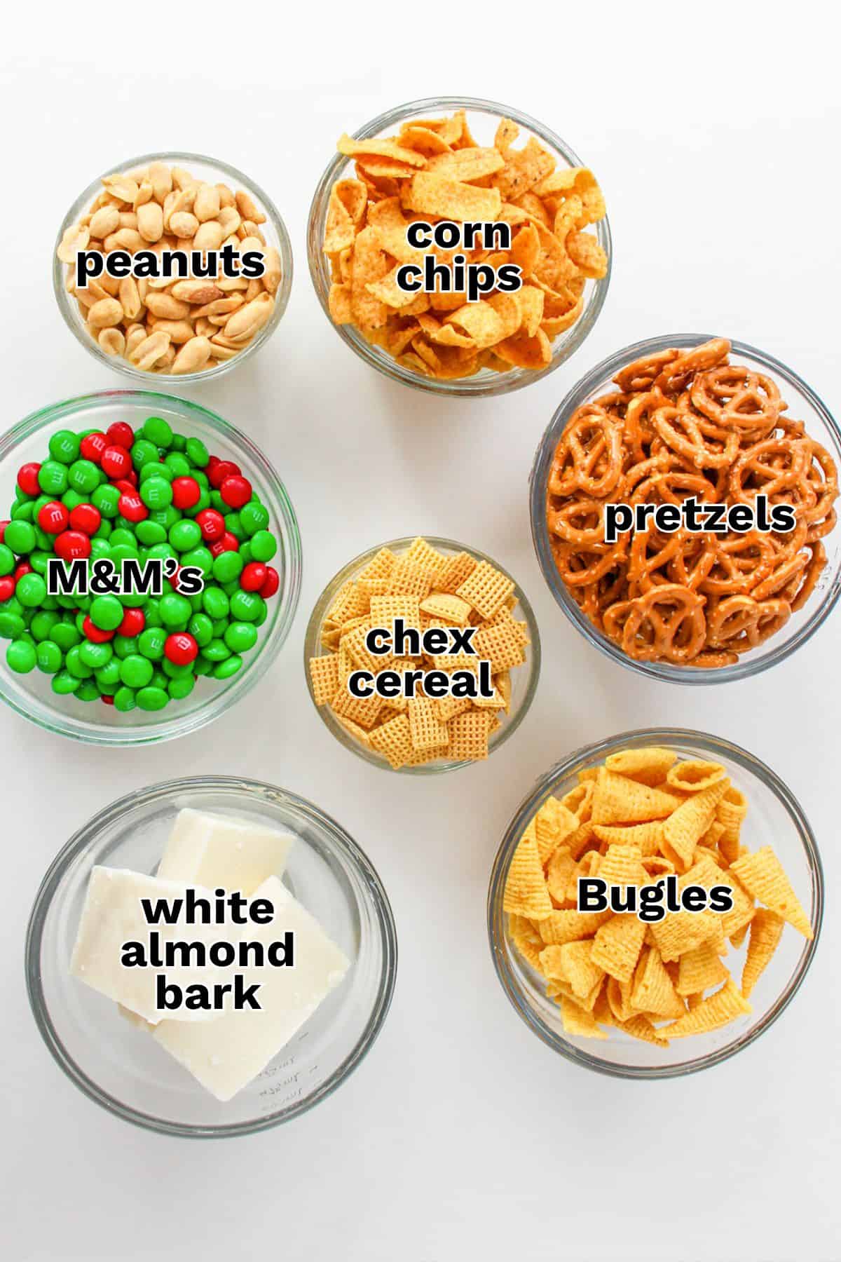 Seven glass bowls on a white surface, each filled with Snack Mix favorites: peanuts, corn chips, pretzels, M&M's, Chex cereal, Bugles, and blocks of white almond bark—perfect for making a classic Christmas White Trash Recipe.